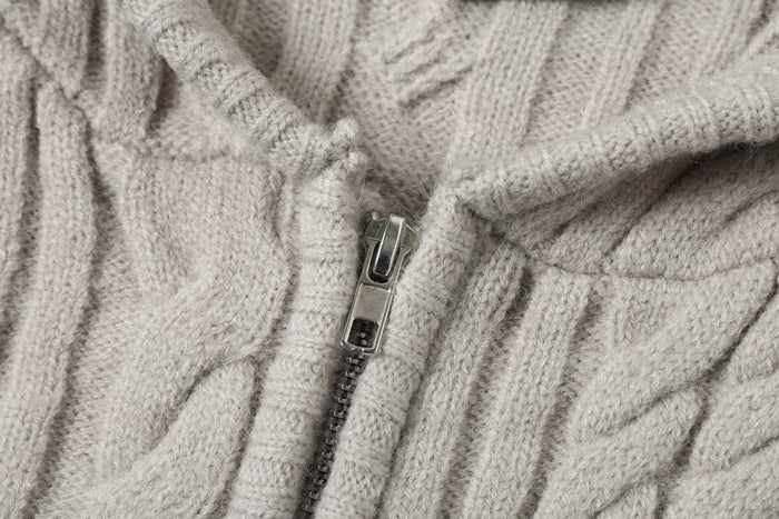 Close-up of a beige knitted hoodie showing the metal zipper, zipper tape, and surrounding cable-knit pattern
