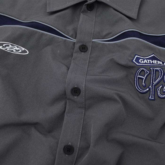 Close-up of gray shirt front showing black buttons, navy-and-white piping, and embroidered chest graphics