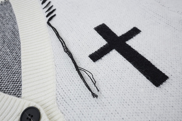 Detail of a black cross motif on white knit fabric beside the V-neck trim and dangling distressed threads