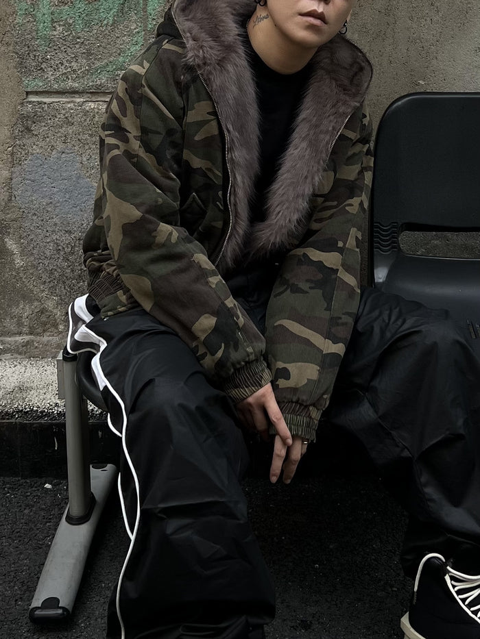 Model seated in a camouflage bomber jacket with a brown faux fur-lined hood and open front