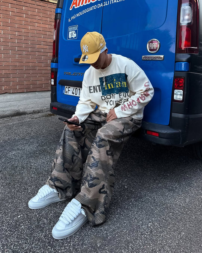 Man seated in cream graphic long sleeve shirt, camo pants, and white sneakers