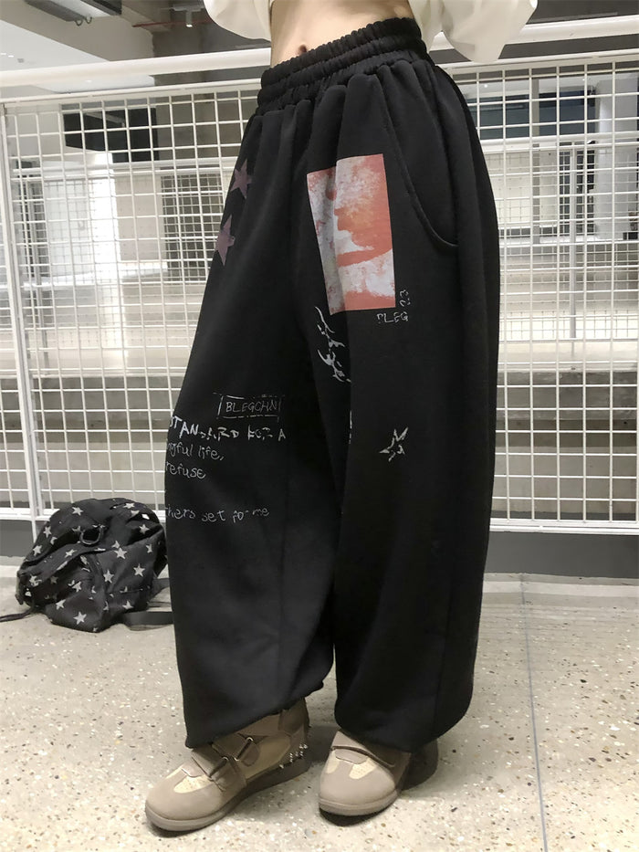 Angled view of black wide-leg sweatpants showing the side pocket, red graphic patch, stars, and printed text