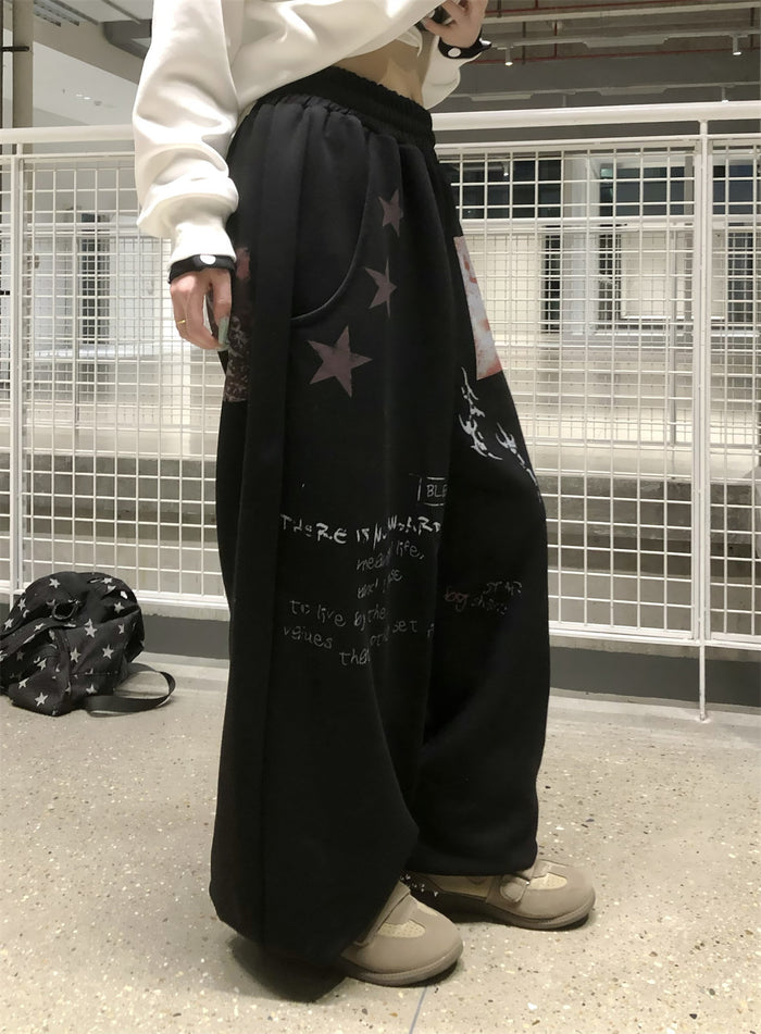 Side view of black wide-leg sweatpants on a model, showing star motifs and layered white graphic lettering