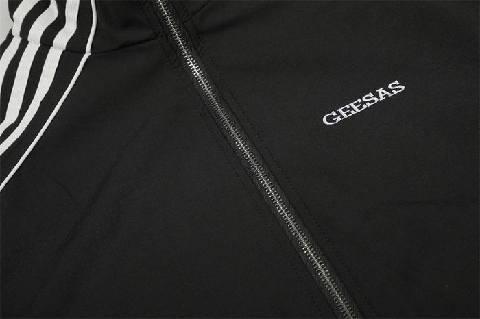 Close-up of the black jacket front showing the metal zipper, white piping, sleeve stripes, and GEESAS logo