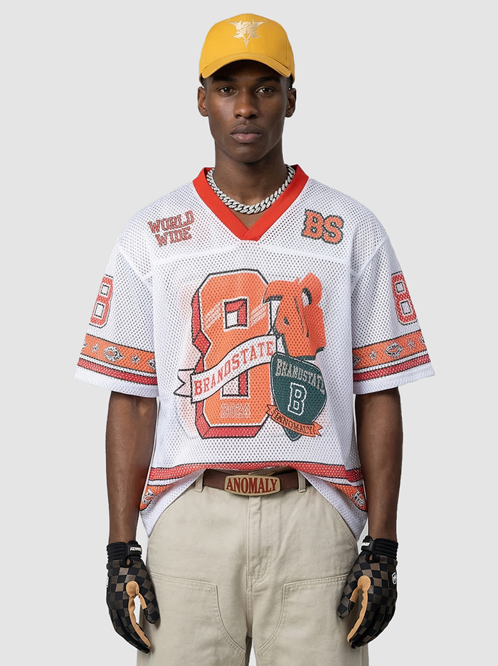 Model wearing a white mesh varsity jersey with orange graphics, red trim, beige cargo pants, and a yellow cap