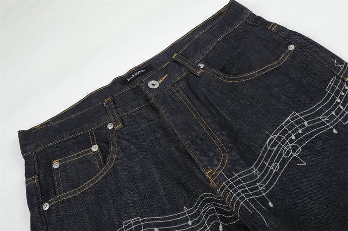 Diagonal front close-up of dark raw denim jeans showing waistband, pockets, and music embroidery