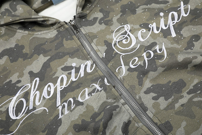 Close-up of gray-green camouflage hoodie front showing the metal zipper, white cursive lettering, and glitter specks